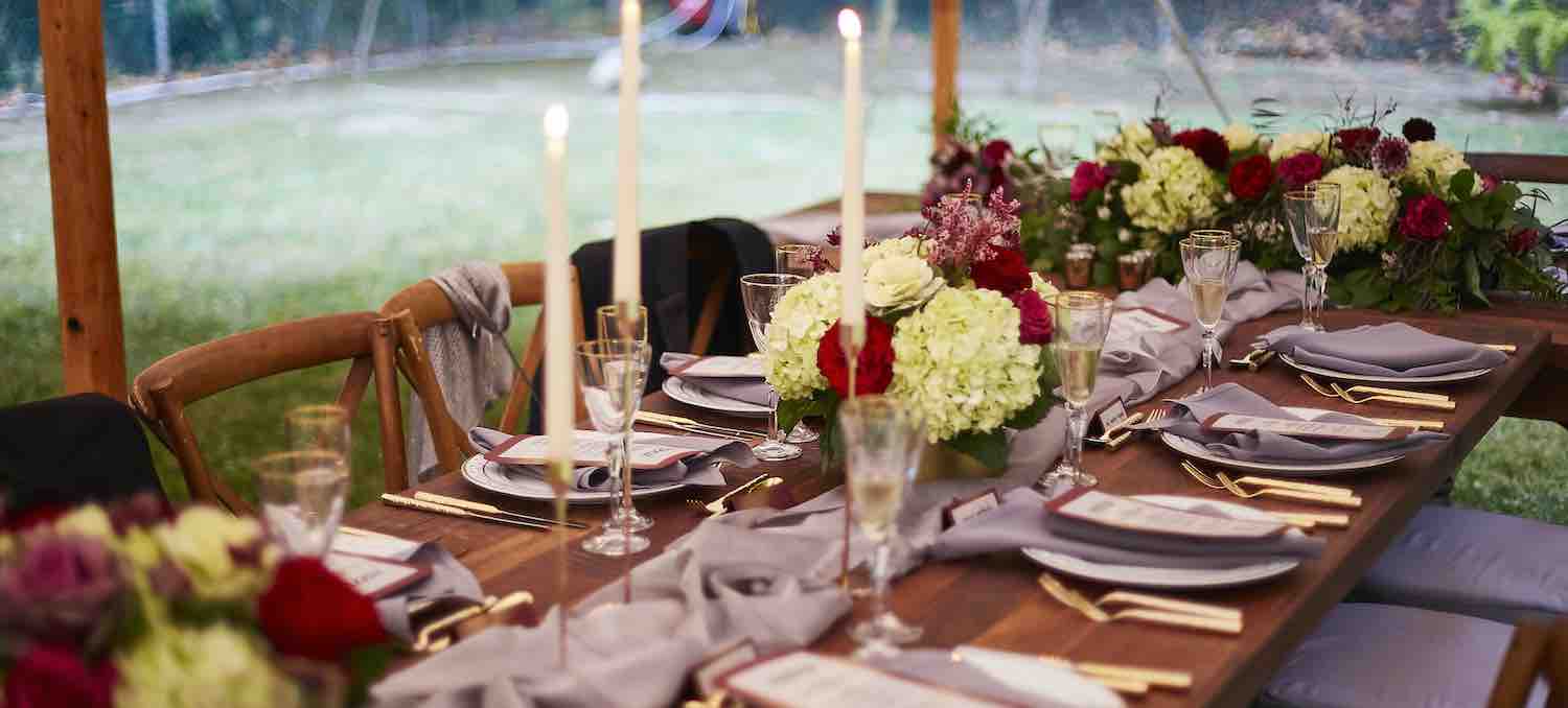Outdoor dining setup under a tent with wooden tables, floral centerpieces, lit candles, and place settings for a formal event.