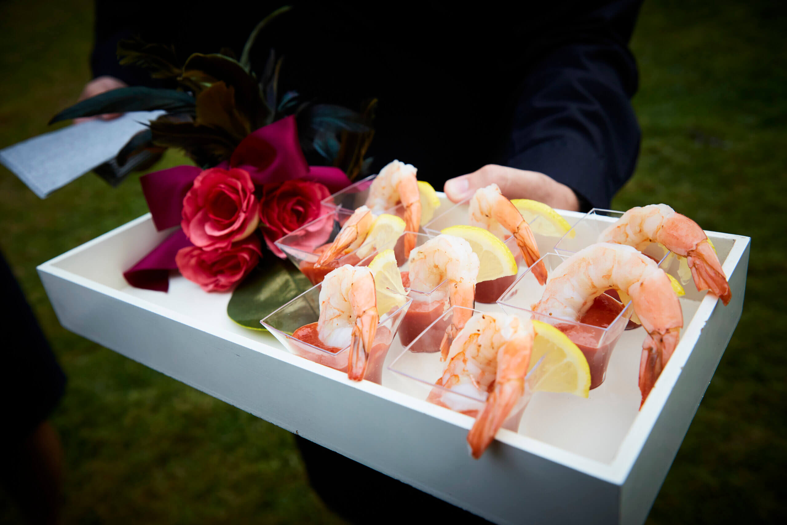 A tray holding shrimp cocktails in clear cups garnished with lemon wedges and served with a side of red roses.