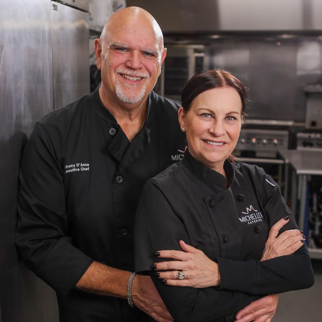 AA6A9172 Two smiling chefs in white uniforms, one woman and one man, standing in a kitchen with arms crossed.